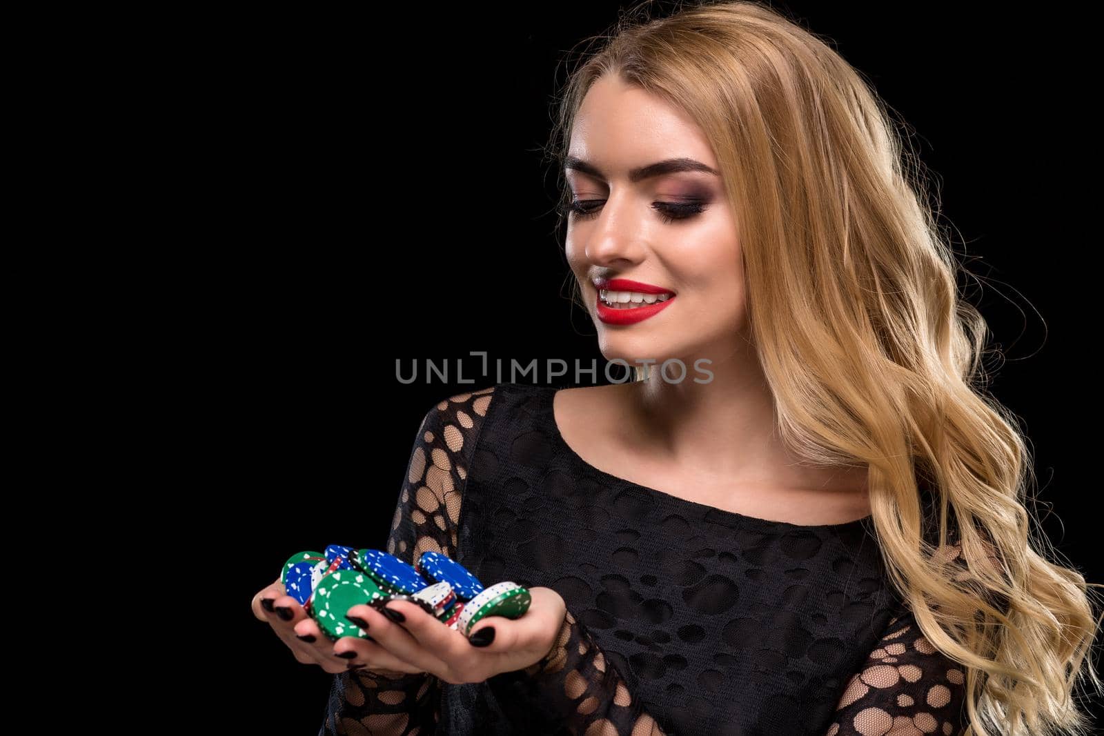 Elegant blonde in a black dress, casino player holding a handful of chips on black background. Poker. Casino. Roulette Blackjack Spin. Caucasian young woman looking at the chips emotions