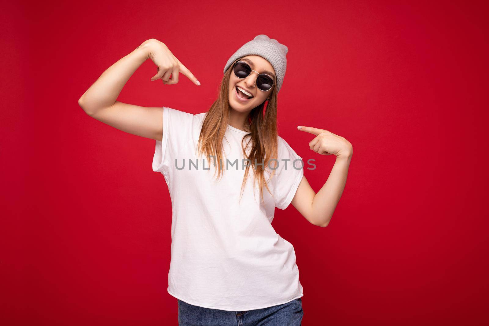 Portrait of young emotional positive happy joyful beautiful dark blonde woman with sincere emotions wearing casual white t-shirt with empty space for mockup gray hat and sunglasses isolated over red background with free space and pointing at herself.