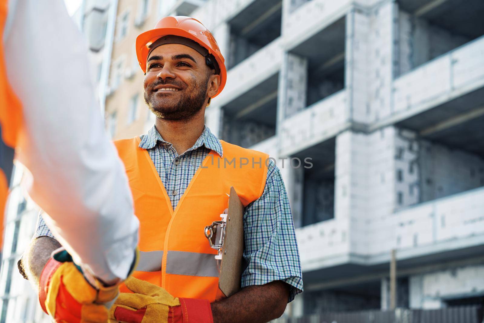 Two men engineers in workwear shaking hands against construction site, close up