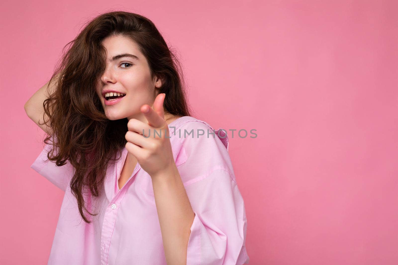 Portrait of beautiful happy smiling positive young curly brunette woman wearing pink shirt and grey hat isolated on pink background with copy space and pointing finger at camera.