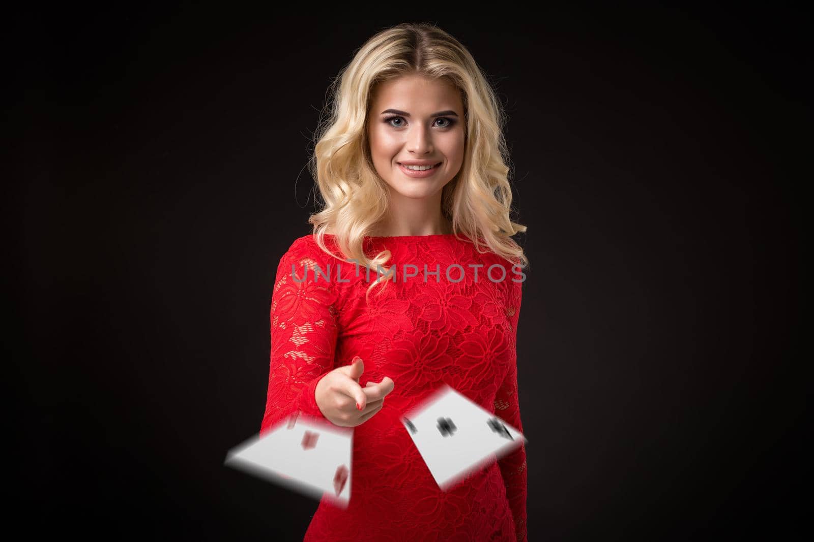 Young beautiful emotional woman throws two aces cards on a black background in the studio. Portrait of a beautiful blonde in a red dress. Poker