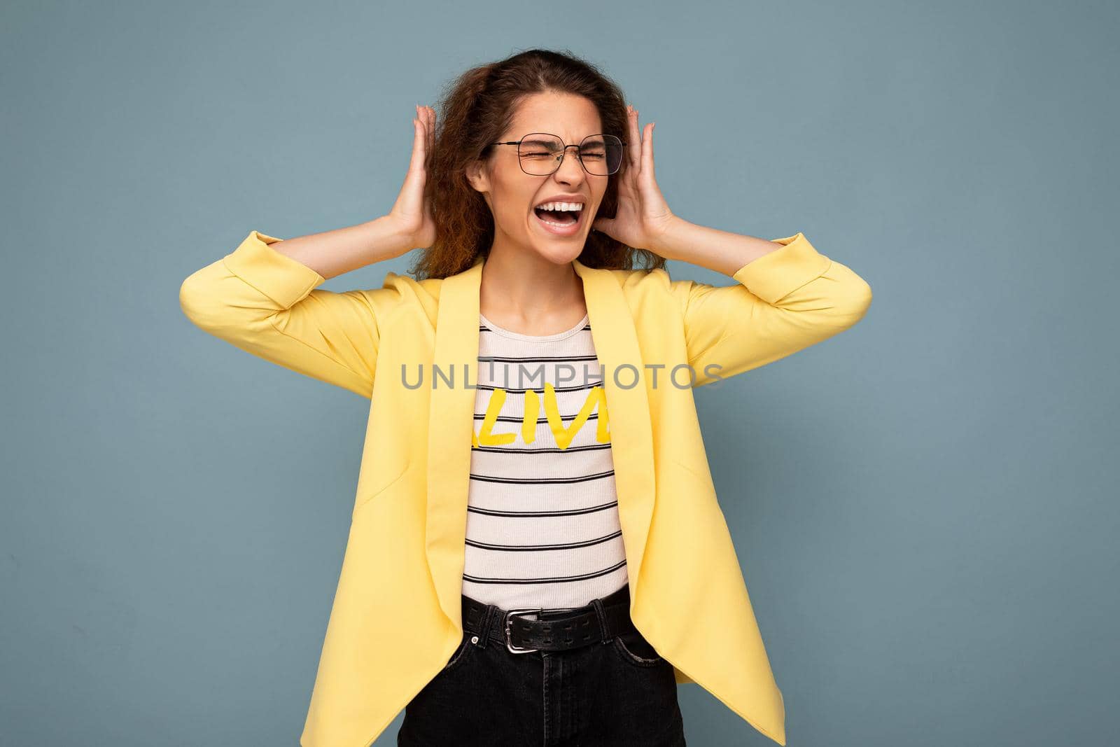 Photo of young emotional angry attractive brunette curly woman with sincere emotions wearing stylish yellow jacket and optical glasses isolated on blue background with empty space and covering ears and shouting.