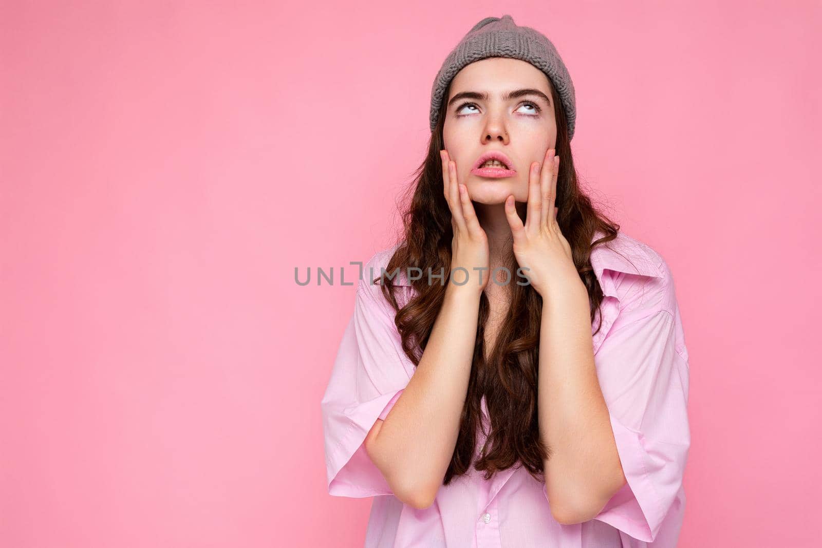 Attractive tired weary young curly brunette woman wearing pink shirt and gray hat isolated on pink background with copy space.