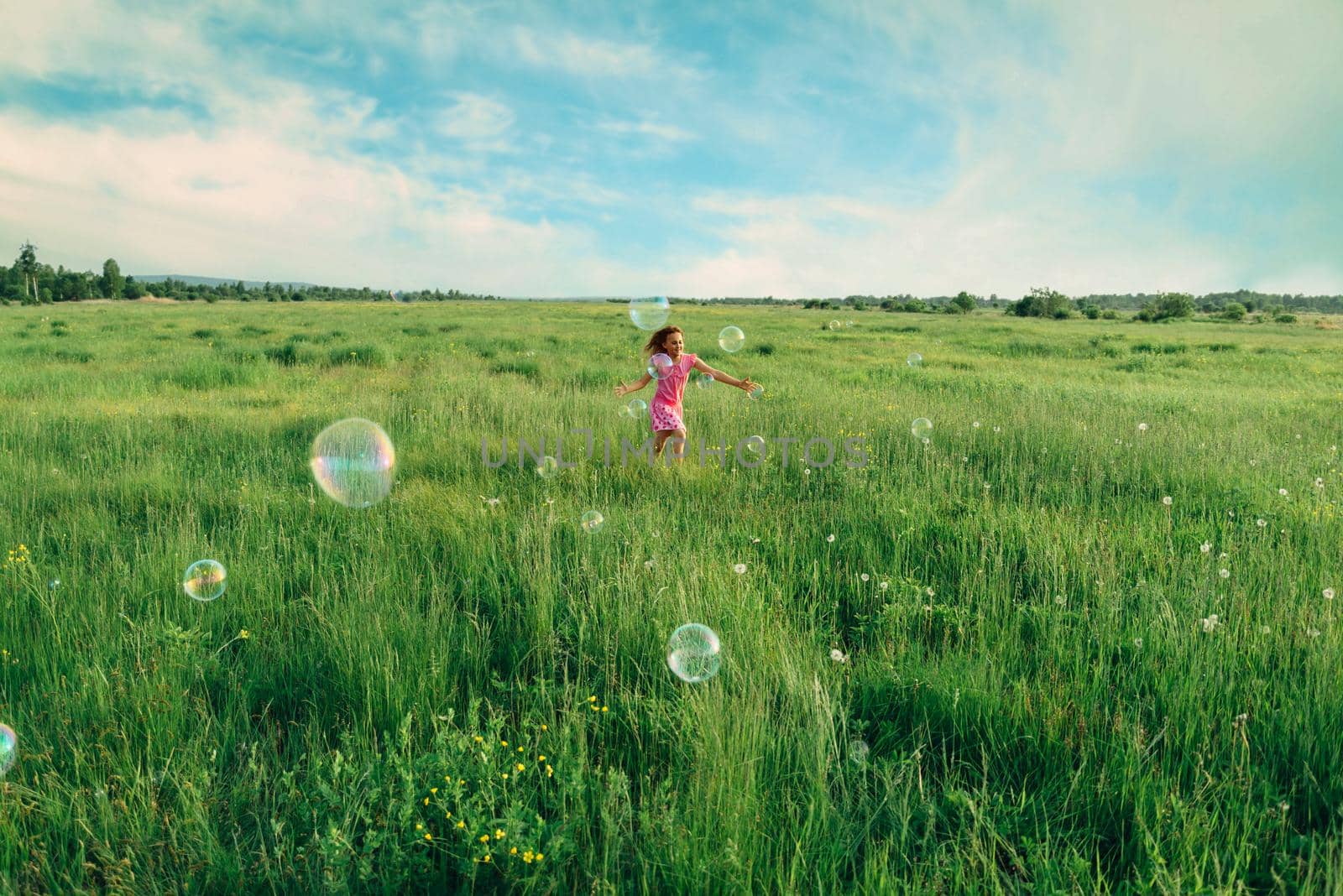 Happy little girl playing among soap bubbles on green meadow in summer
