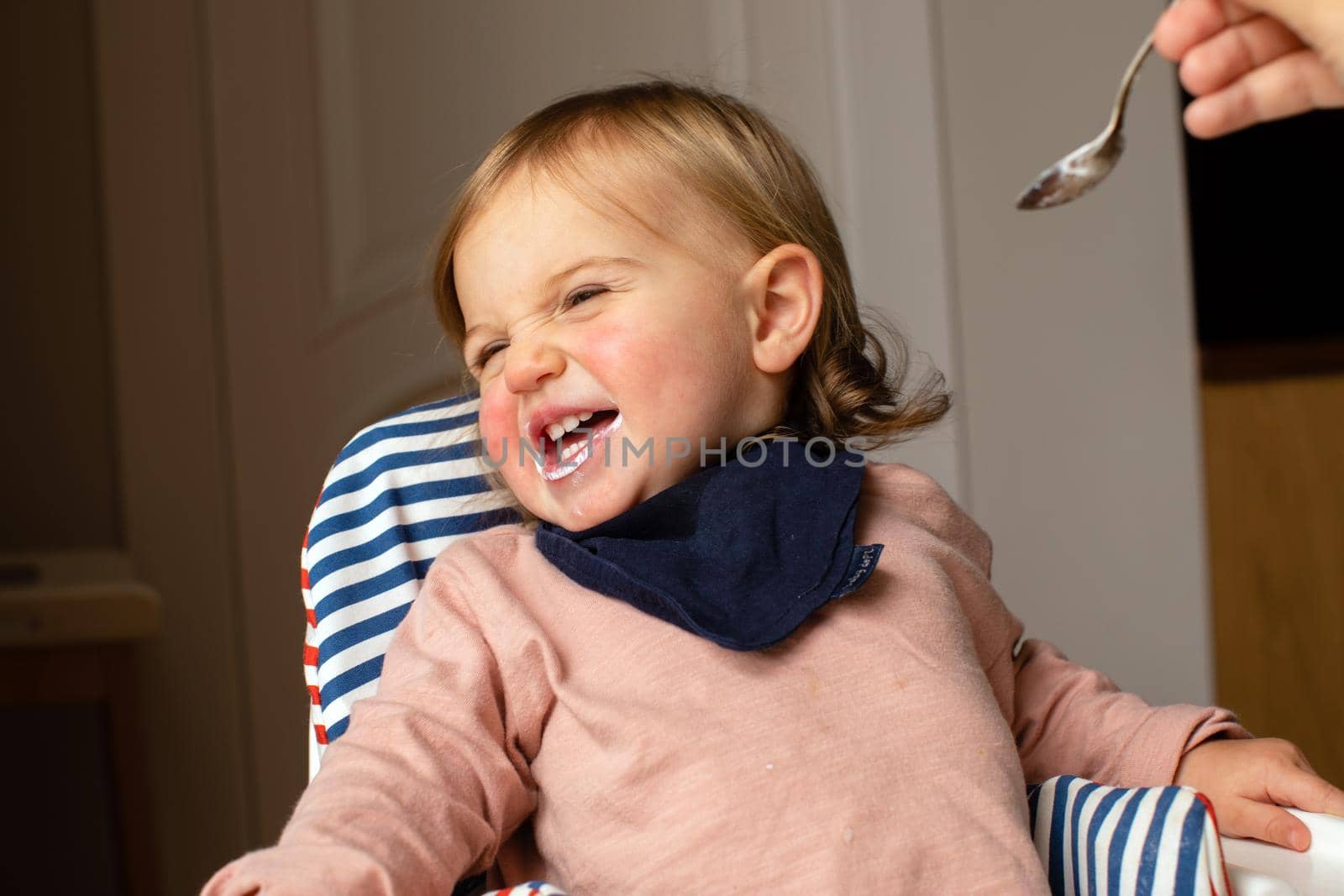 Adorable child smiling and grimacing tossing head in high chair while crop person feeding with spoon in room