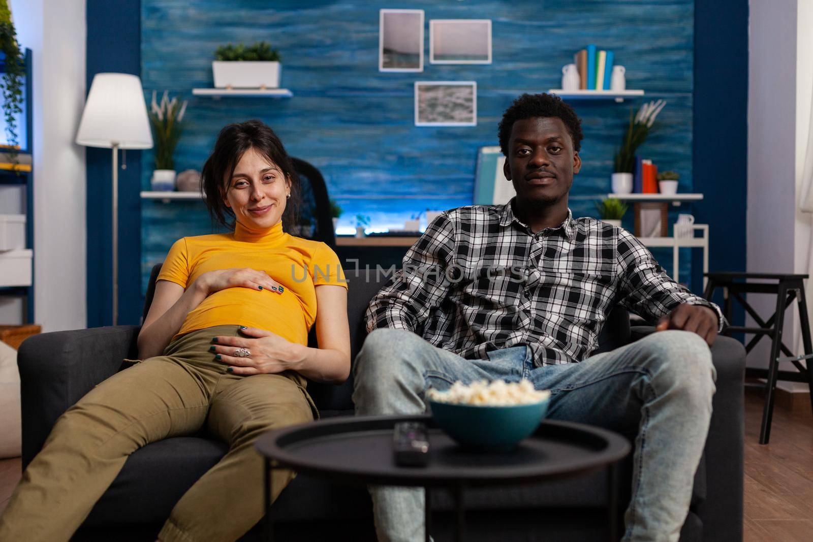 POV of interracial people expecting baby watching television at home. Multi ethnic couple with pregnancy looking at camera and enjoying movie. Pregnant woman holding hand on baby bump