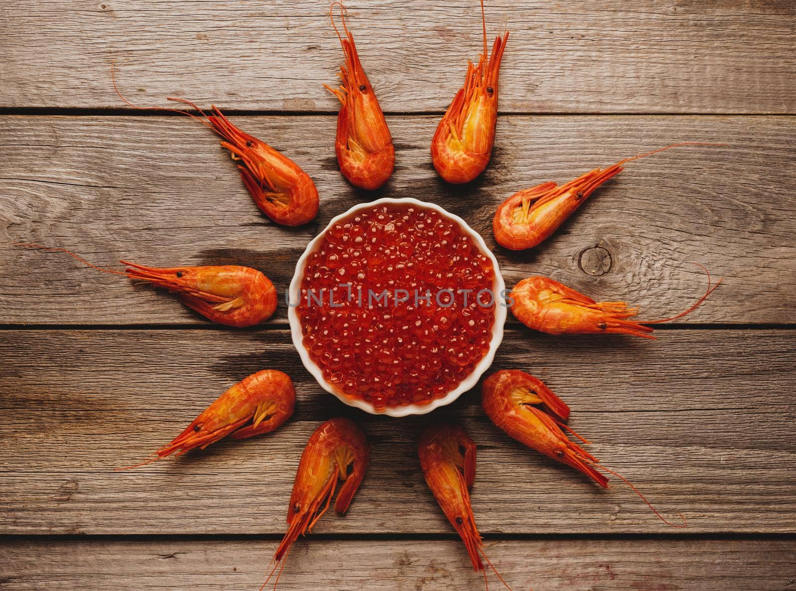 Seafood: boiled shrimps around bowl with red caviar on wooden background, top view