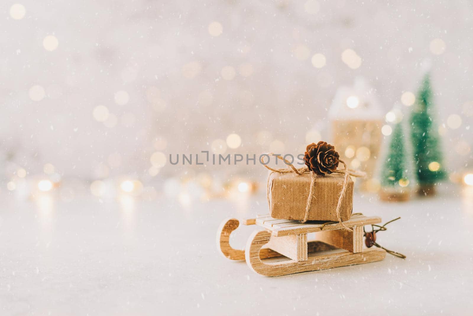 Eco Natural Christmas gift on toy wooden sled on white background with glowing light bokeh, copy space. New year card. Xmas decoration with house, Christmas tree and snow