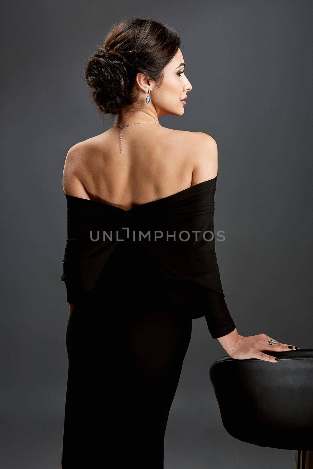 Beautiful woman standing in a black dress over gray background. A woman is standing back.
