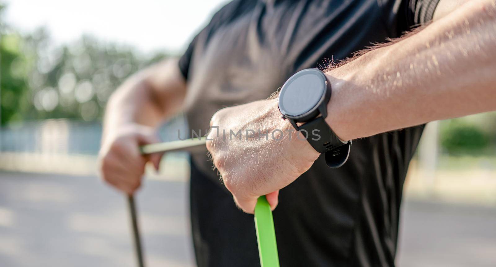 Sportive man guy training with elastic rubber band at the stadium outdoors with sunlight. Athlete male person during workout with additional sport equipmant