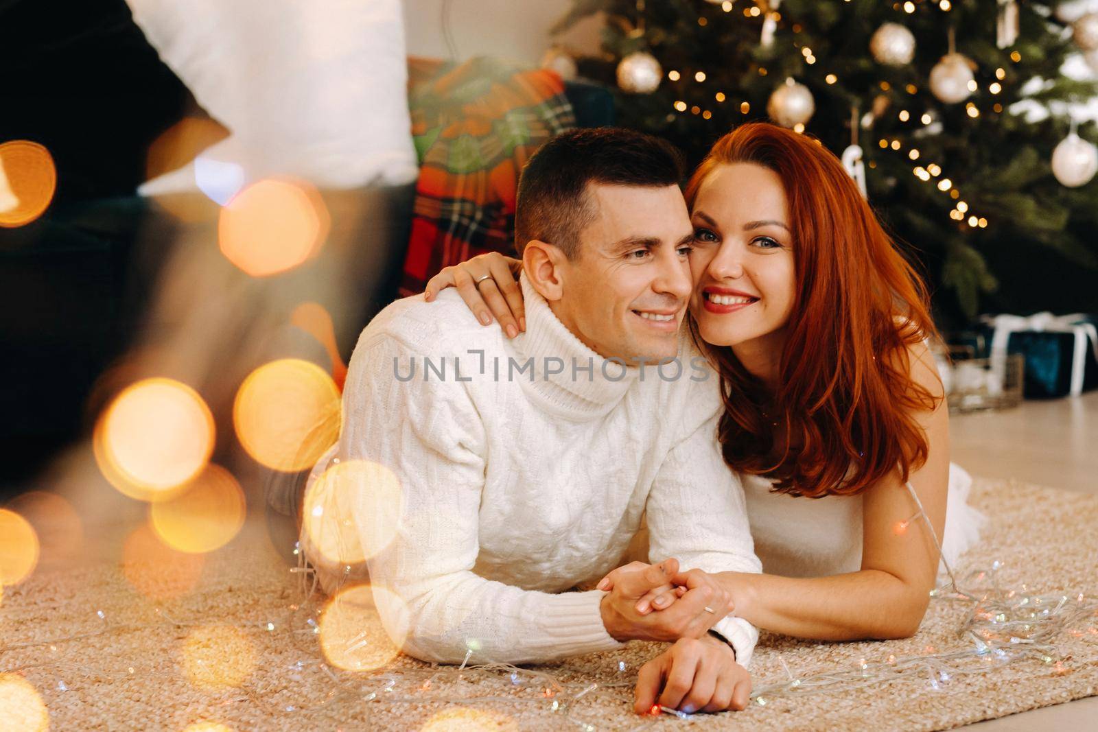 a happy married couple is lying on the floor near the Christmas tree at home by Lobachad