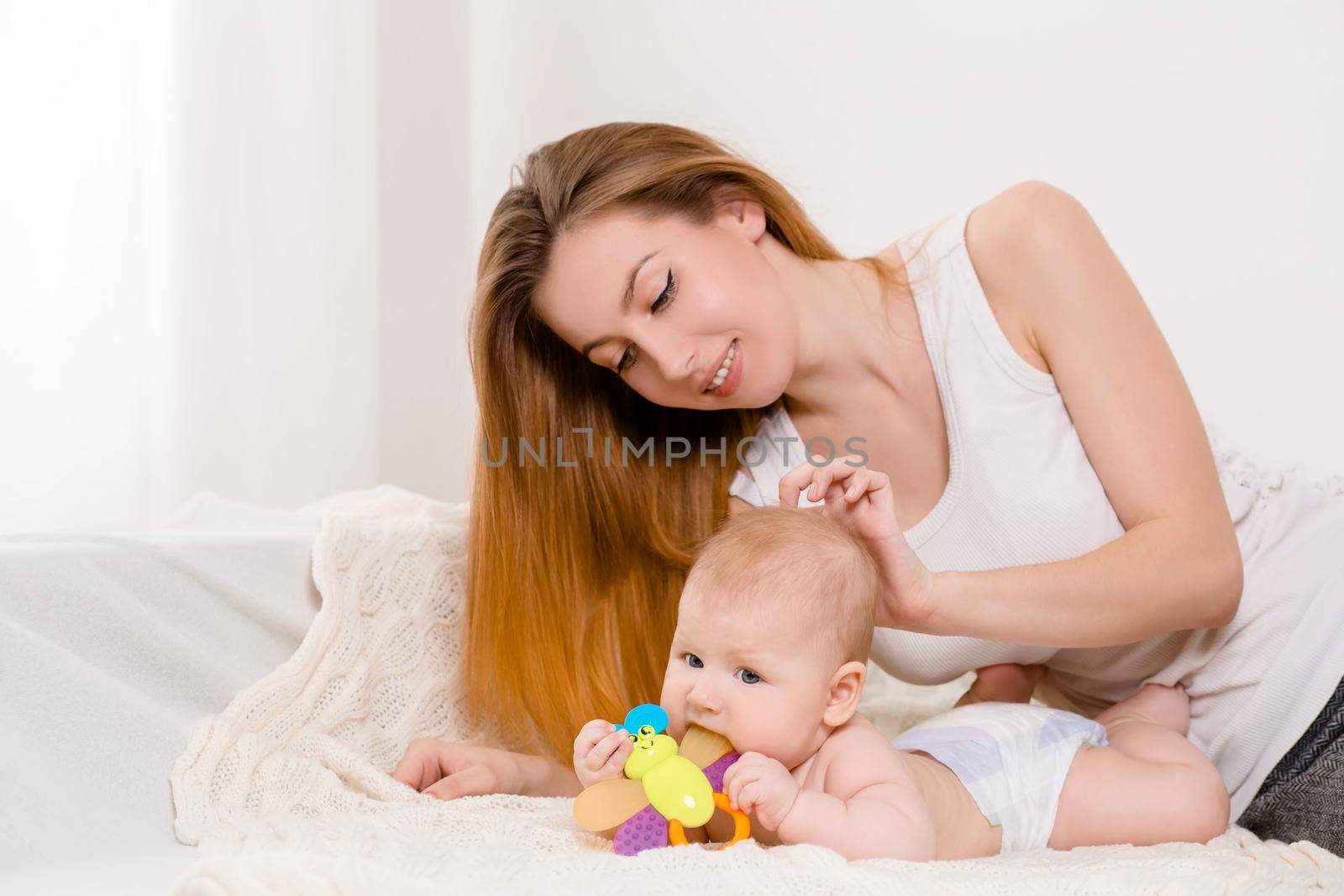 Mother and child on a white bed. Mom and baby girl in diaper playing in sunny bedroom. Parent and little kid relaxing at home. Family having fun together. Bedding and textile for infant nursery.