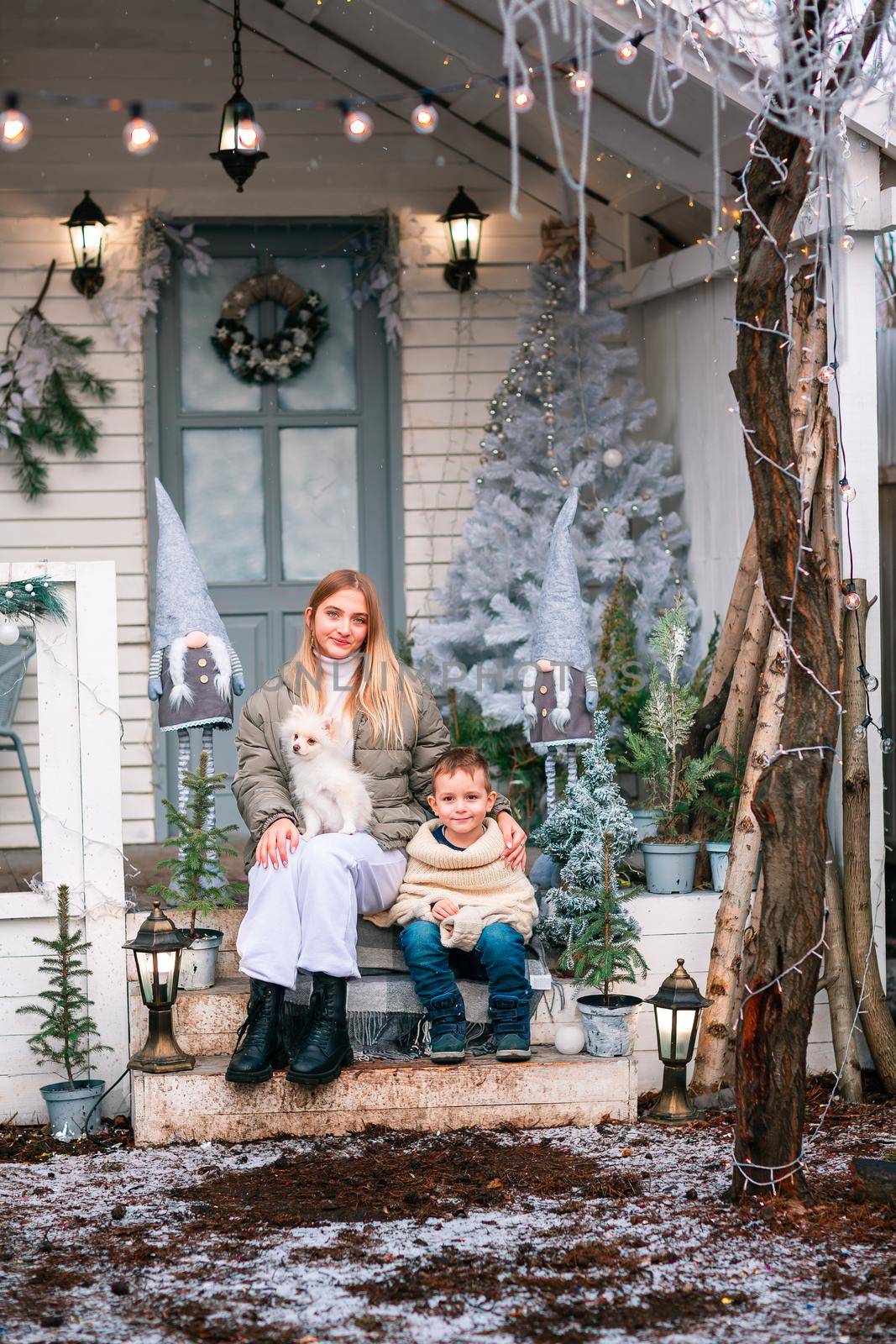 Happy little kids with white little puppy sitting on the porch of the Christmas decorated house, snowing outdoor. Happy New Year and Merry Christmas. Magic winter