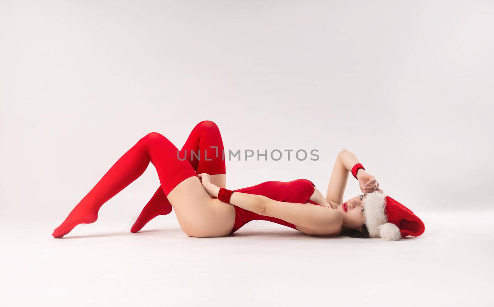 woman in a red Santa hat with Christmas gifts on the floor poses sexually with a white background