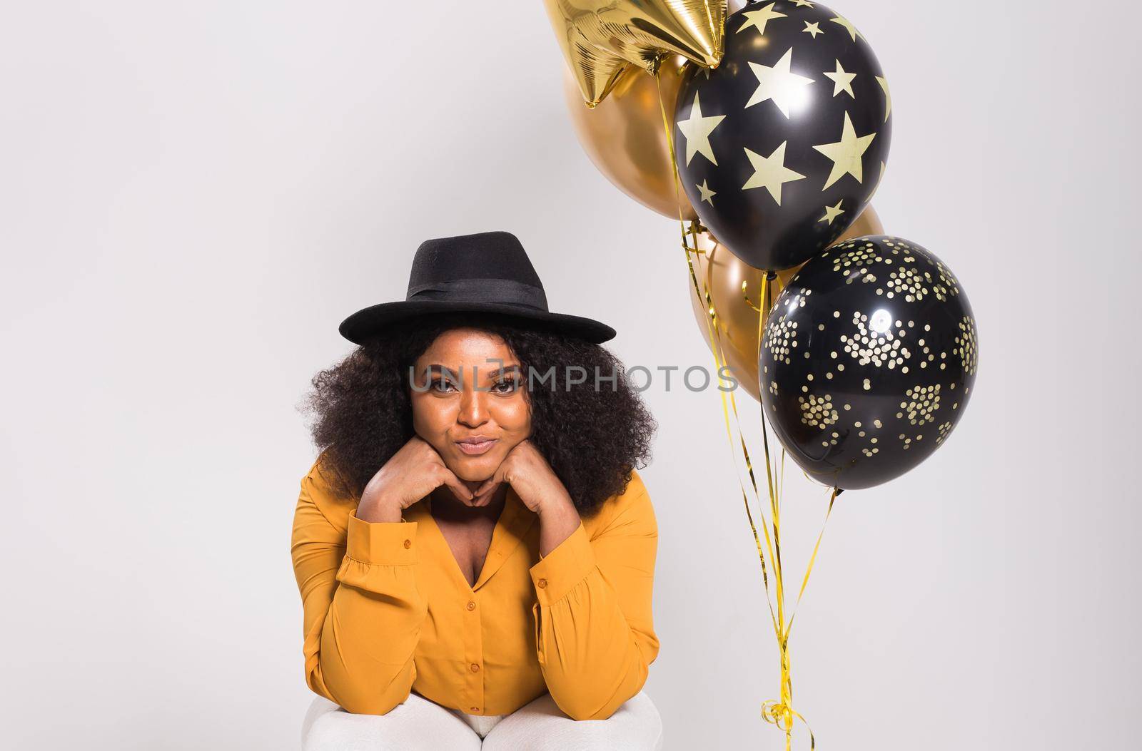 Portrait of smiling young African-American adult woman looking sweet on yellow background holding balloons.
