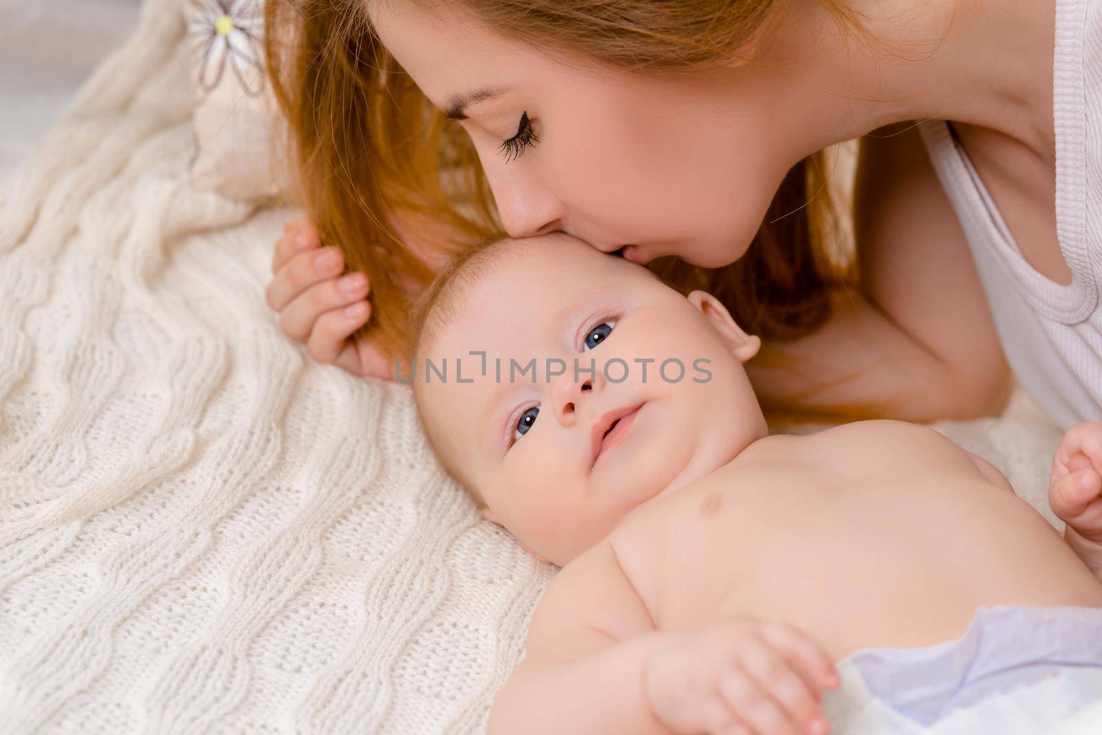 Mother and child on a white bed. Mom and baby girl in diaper playing in sunny bedroom. Parent and little kid relaxing at home. Family having fun together. Bedding and textile for infant nursery.