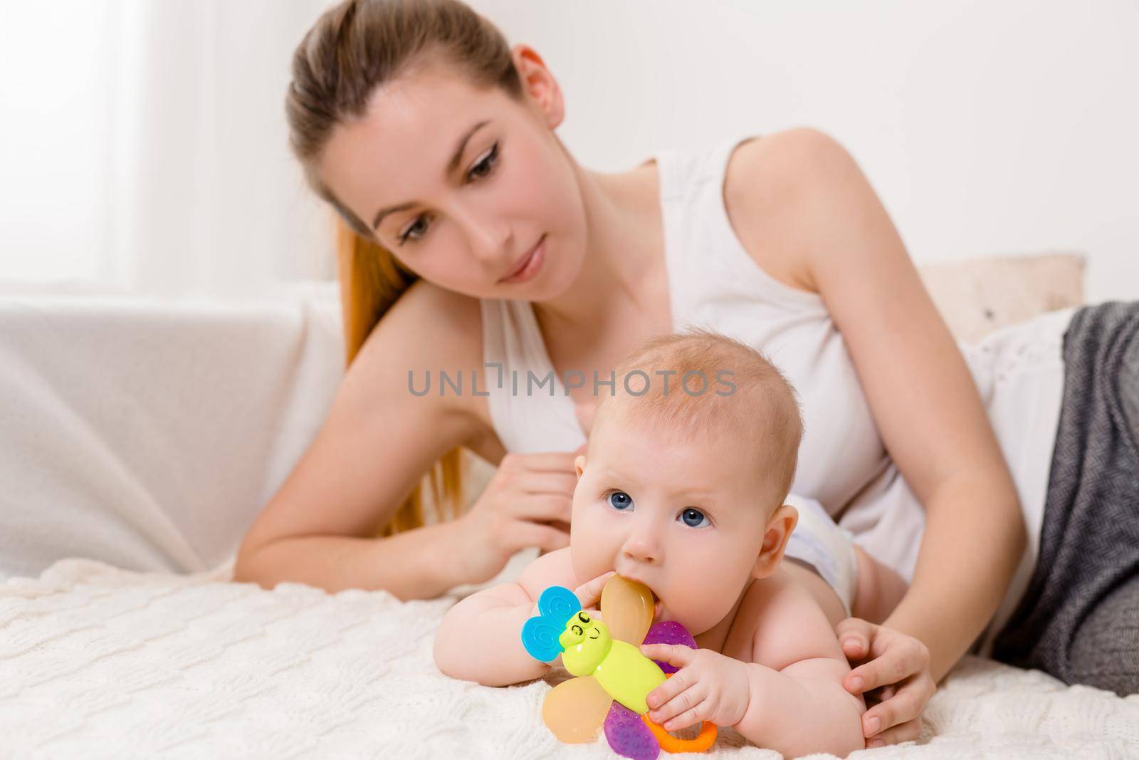 Mother and child on a white bed. Mom and baby girl in diaper playing in sunny bedroom. Parent and little kid relaxing at home. Family having fun together. Bedding and textile for infant nursery.