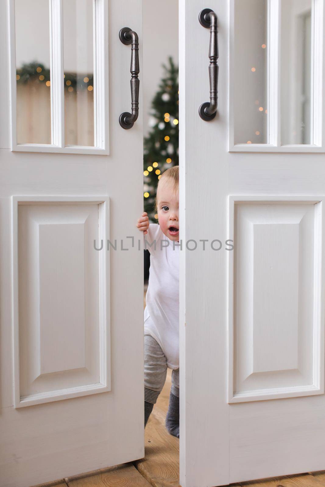 Adorable infant standing in doors looking at camera opens the door and is surprised on background of Christmas tree