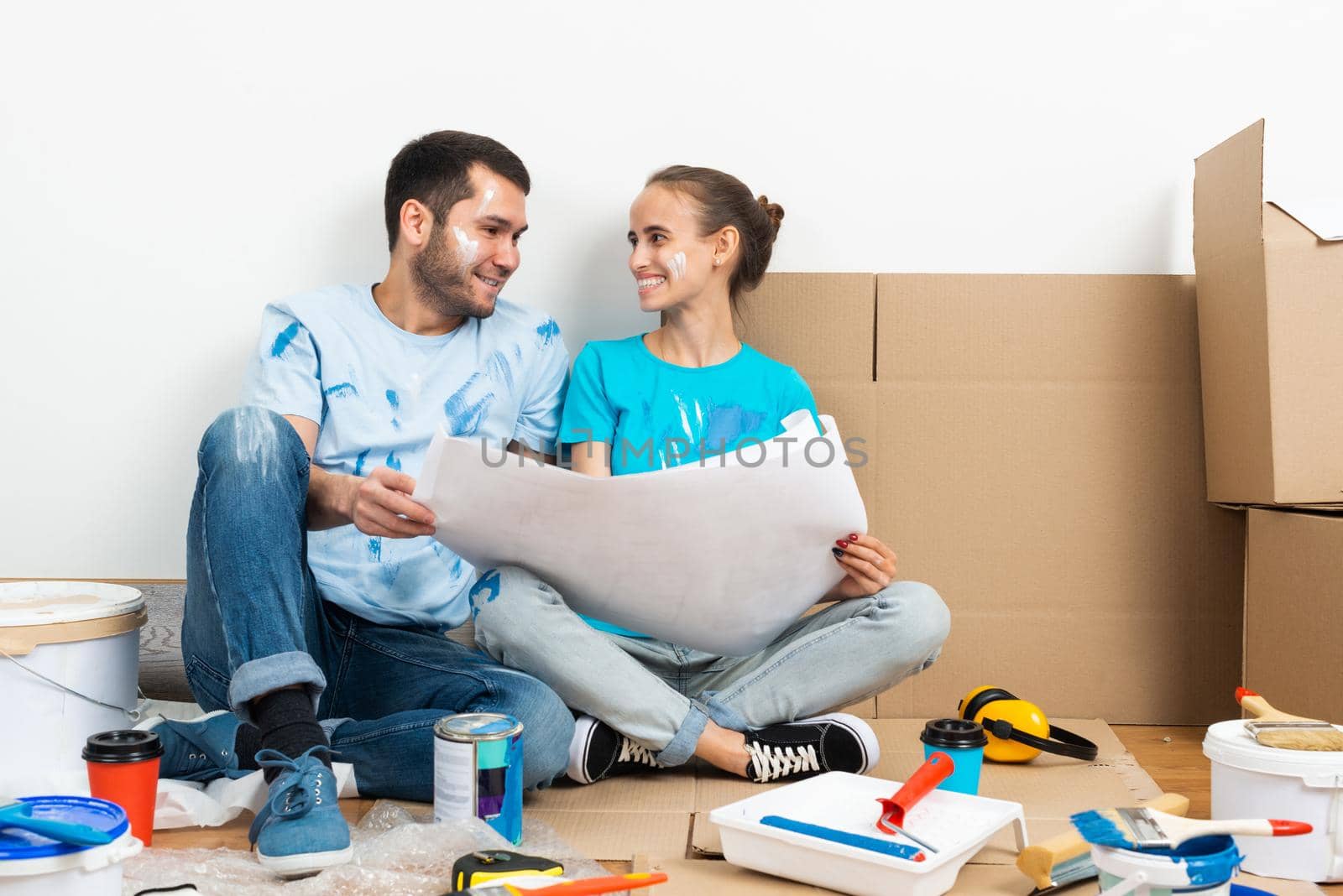 Couple moving into new house concept. Smiling young man and woman together sitting on floor among cardboard boxes. House remodeling and interior renovation. People looking at blueprint at home.