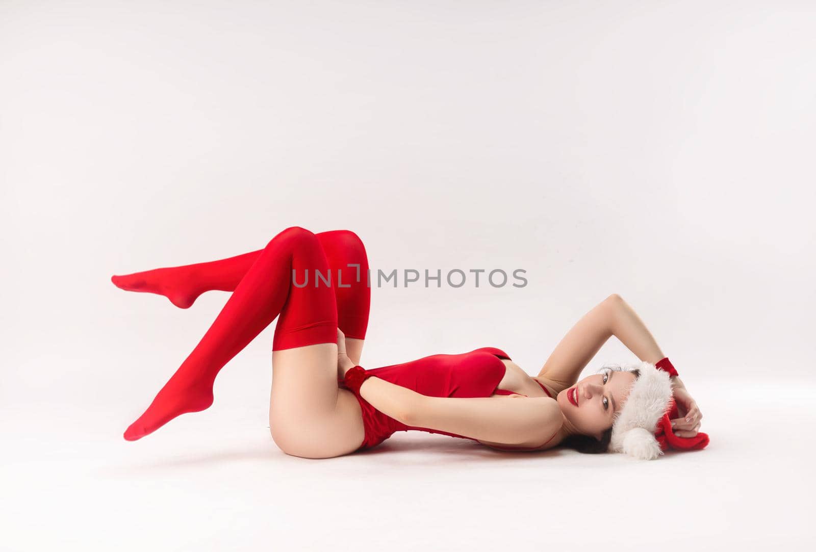 woman in a red Santa hat with Christmas gifts on the floor poses sexually with a white background