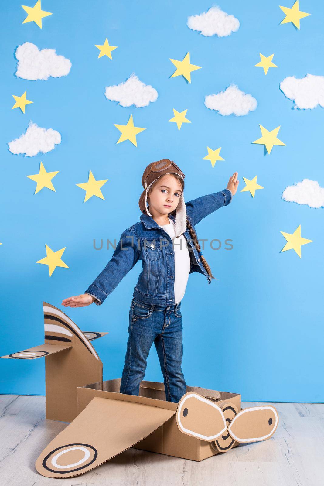 Little child girl in a pilot's costume is playing and dreaming of flying over the clouds. Portrait of funny kid on a background of bright blue wall with yellow stars and white clouds