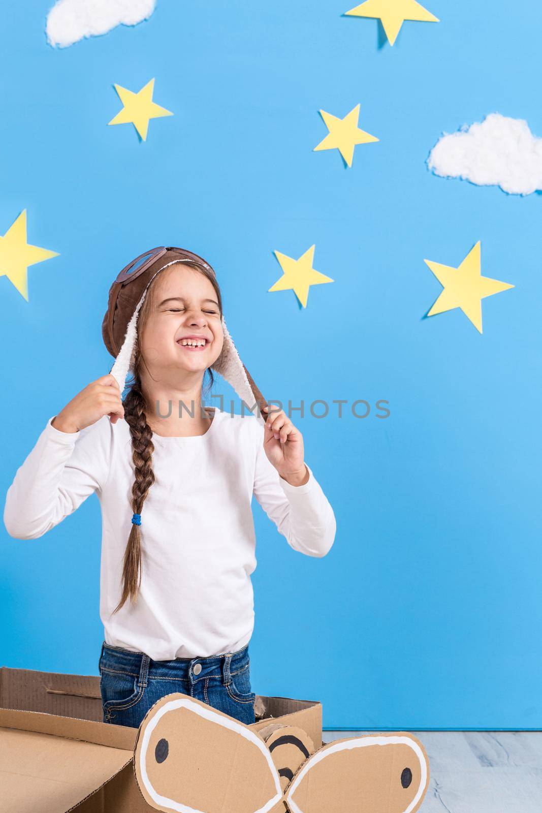 Little child girl in a pilot's costume is playing and dreaming of flying over the clouds. Portrait of funny kid on a background of bright blue wall with yellow stars and white clouds