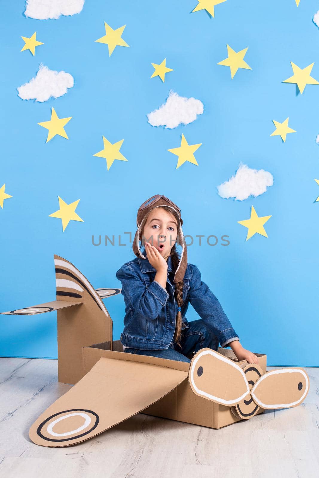 Little dreamer girl playing with a cardboard airplane at the studio with blue sky and white clouds background. Childhood. Fantasy, imagination.