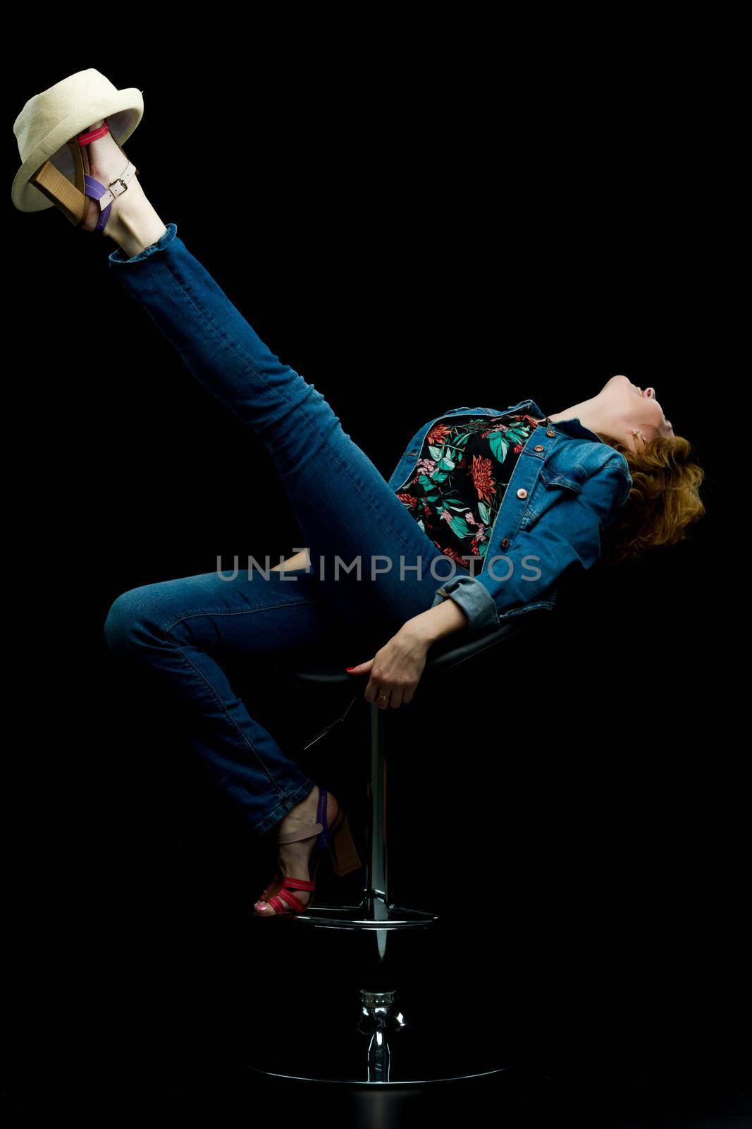 Beautiful young woman posing studio in a bar stool, she put a hat on her leg. Photo joke. On black background