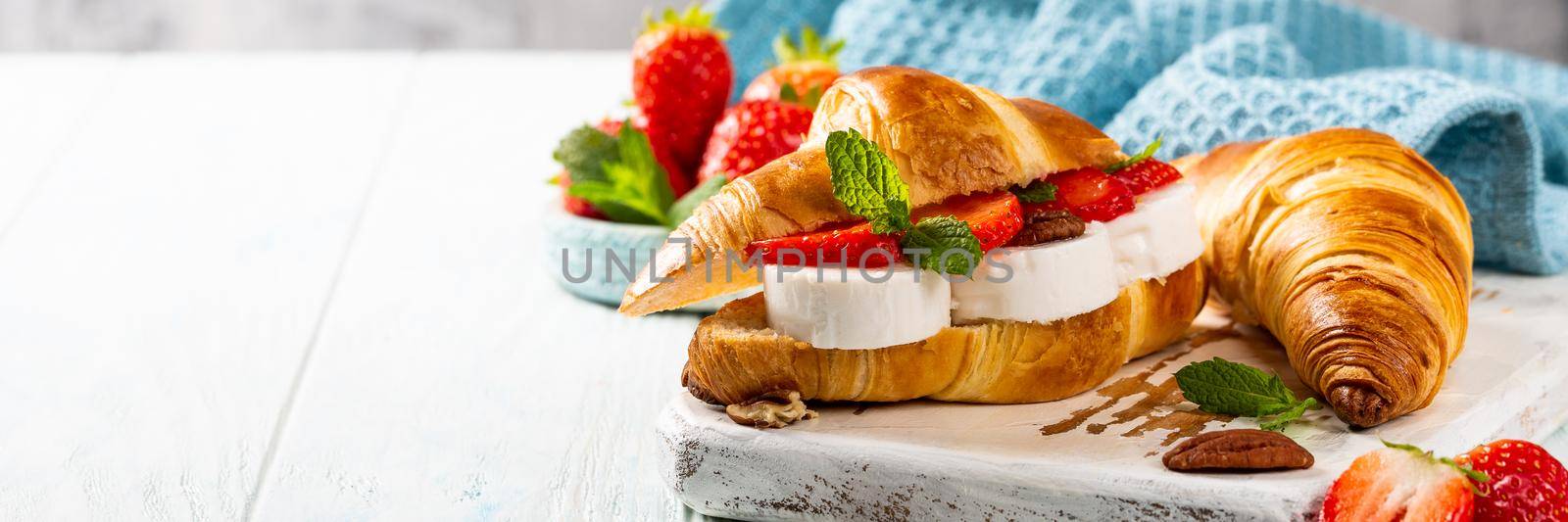 Helthy breakfast with croissant with goat cheese, strawberries, honey and pecans on wooden cutting board with copy space. Banner.