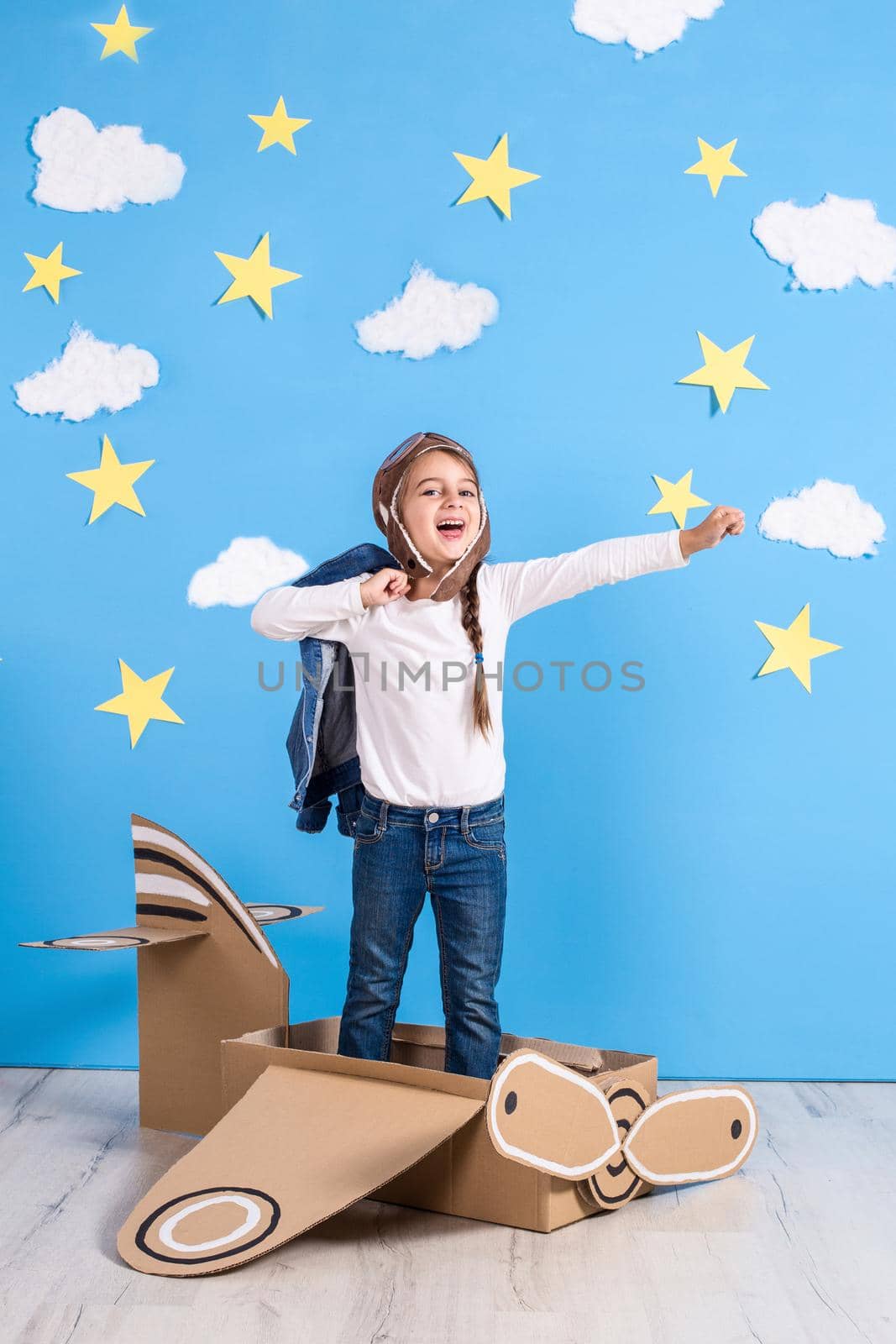 Little dreamer girl playing with a cardboard airplane at the studio with blue sky and white clouds background. Childhood. Fantasy, imagination.