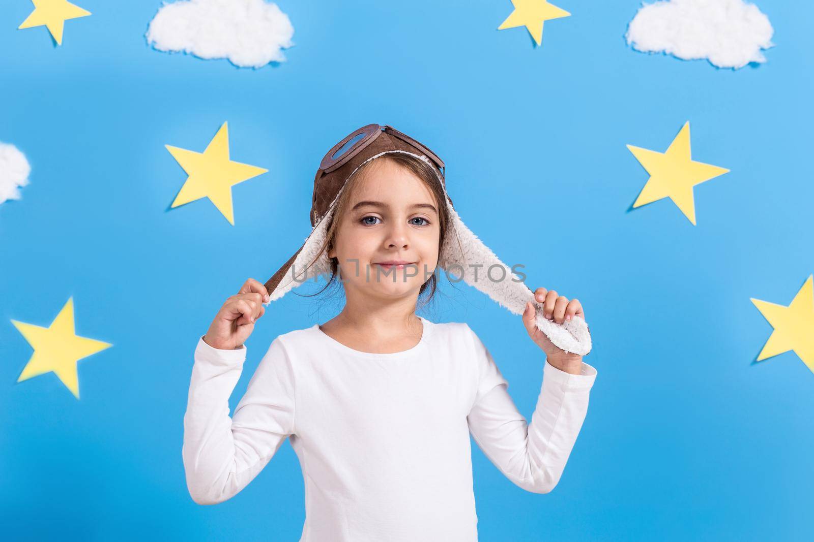 Little happy girl in aviator's cap smiling and playing, in white shirt at the studio. Blue sky, white clouds and stars background. Childhood. Fantasy, imagination.