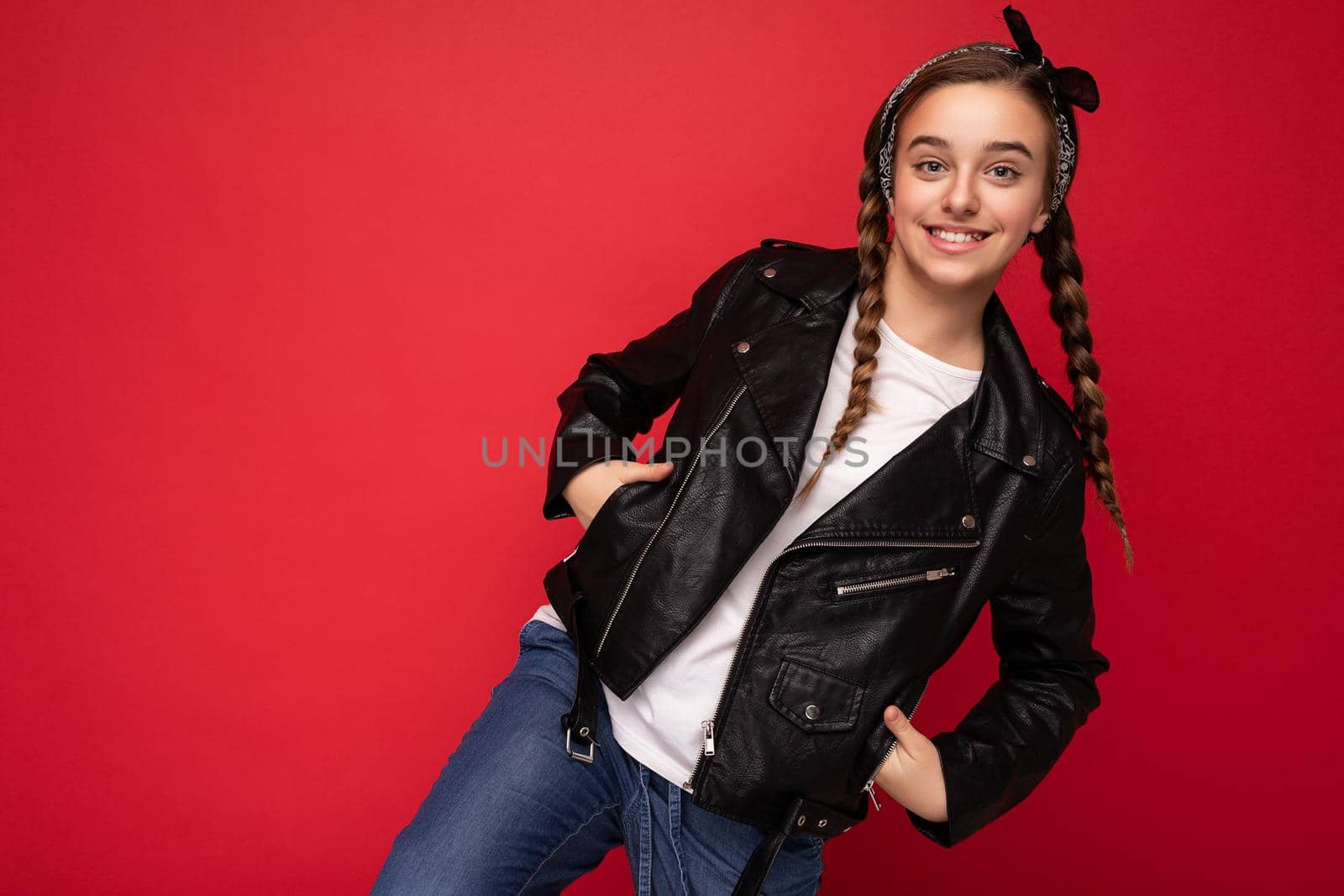 Photo shot of beautiful happy smiling brunette little girl with pigtails wearing trendy black leather jacket and white t-shirt standing isolated over red background wall looking at camera. Copy space