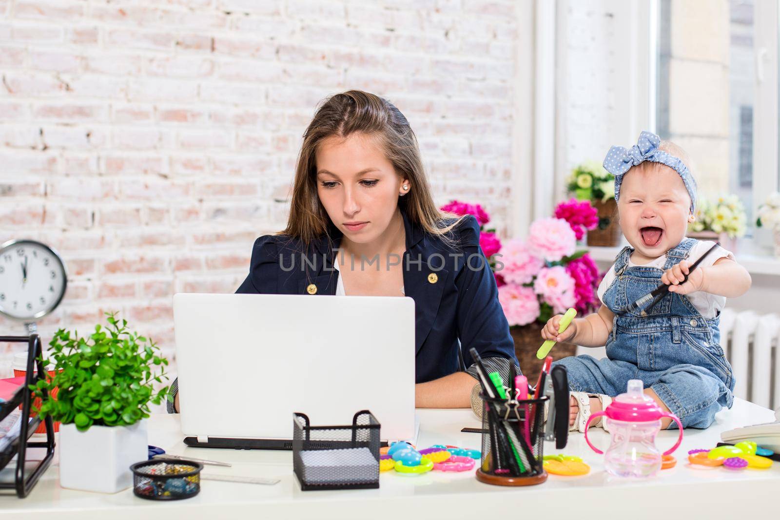 Cheerful young beautiful businesswoman looking at laptop while sitting at her working place with her little daughter by nazarovsergey