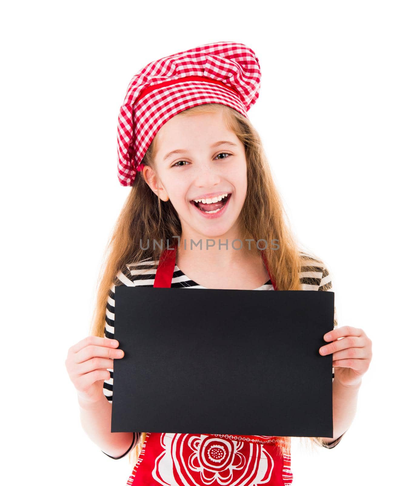 Cute little girl in red kitchen uniform holds horizontal blank black sheet and smiles isolated on white background. Copyspace, space for your text, recipe, advertising, menu