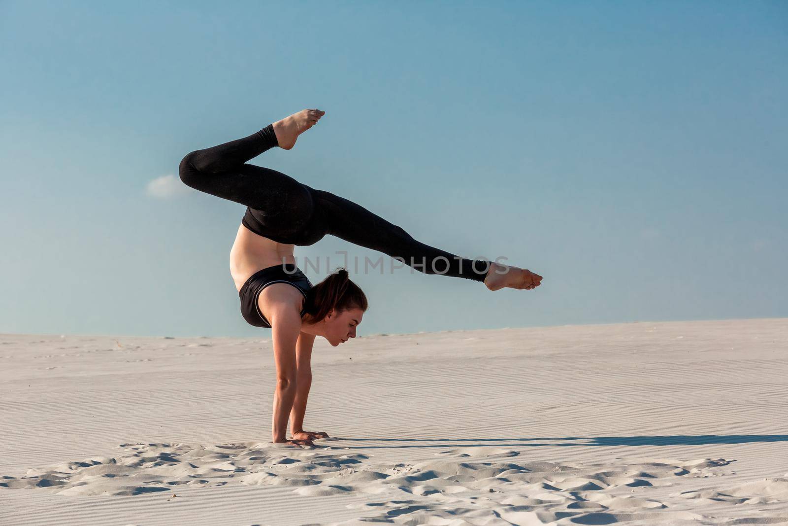 Young woman practicing inversion balancing yoga pose handstand on beach with white sand and bright blue sky