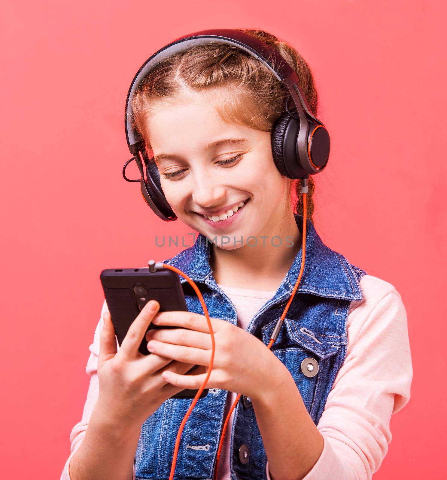 Happy teenage girl listening to music in big headphones while playing with smartphone on pink background