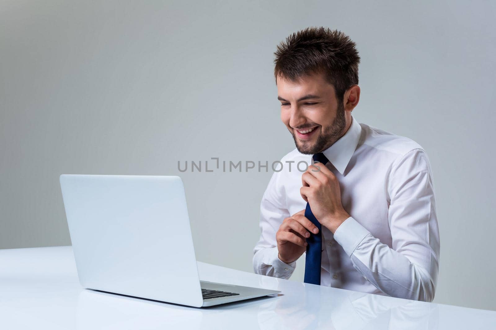 young man smiles and straightens his tie using laptop computer sitting at the table. Office clothing