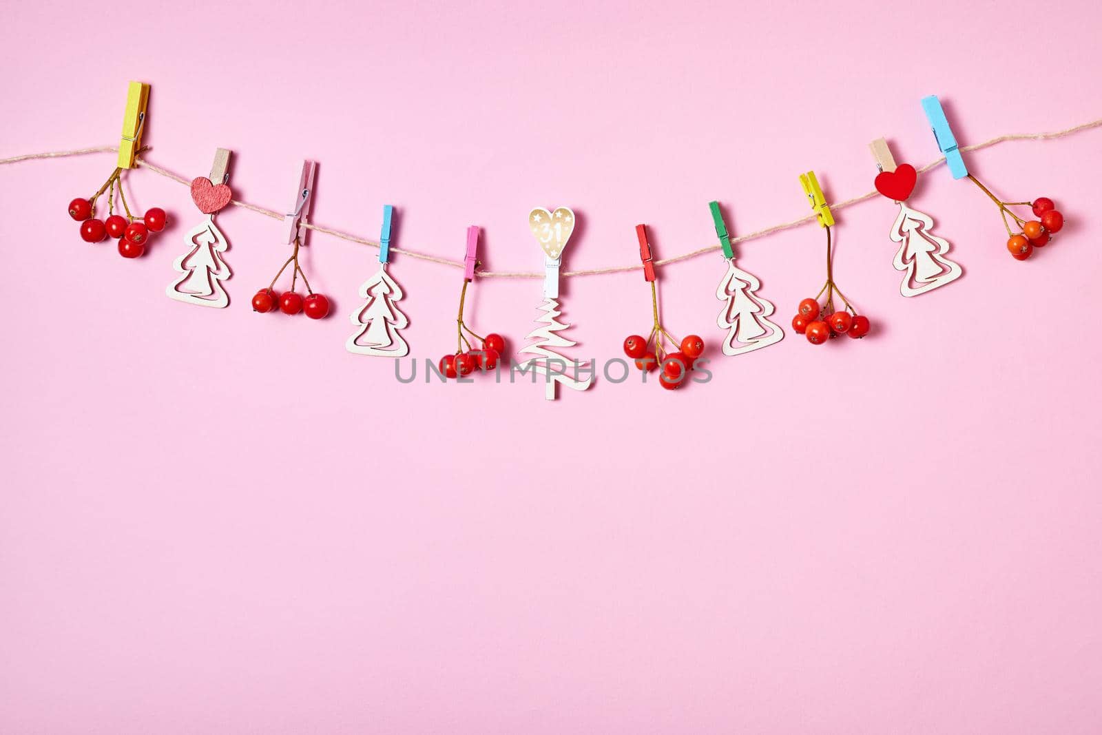 Figurines of wooden Christmas trees and rowan berries are hung with clothespins on a rope on a pink background by marketlan