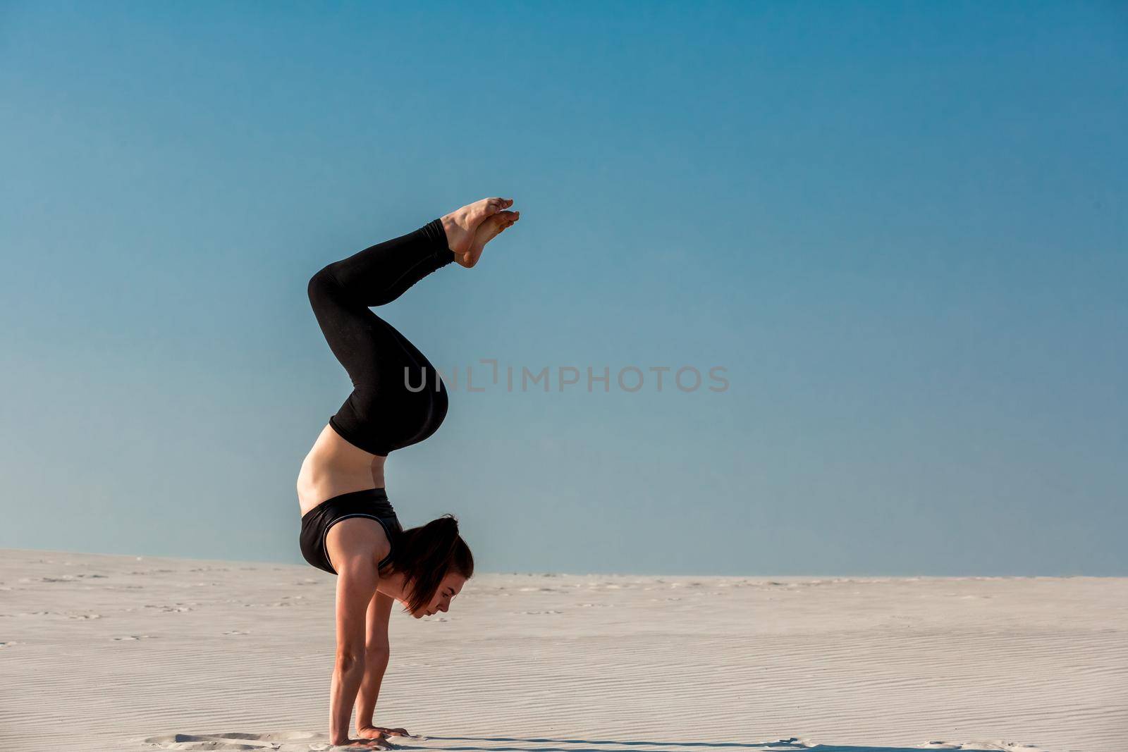 Young woman practicing handstand on beach with white sand and bright blue sky by nazarovsergey