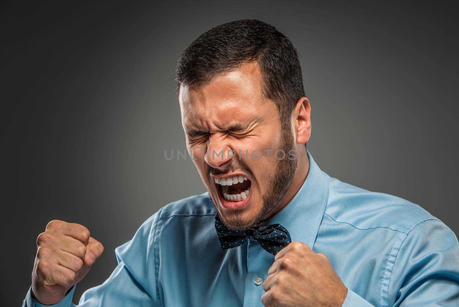 Portrait of angry upset young man in blue shirt and butterfly tie with fists up yelling isolated on gray studio background. Negative human emotion, facial expression. Closeup
