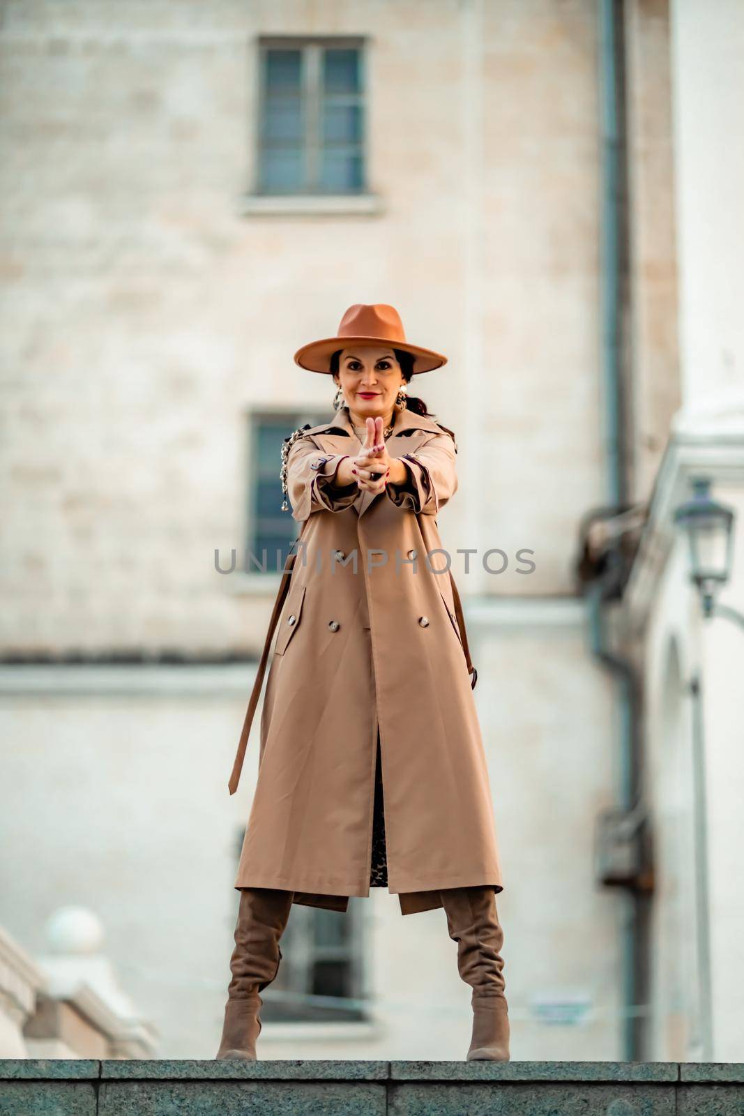 Outdoor fashion portrait of young elegant fashionable brunette woman, model in stylish hat, choker and light raincoat posing at sunset in European city