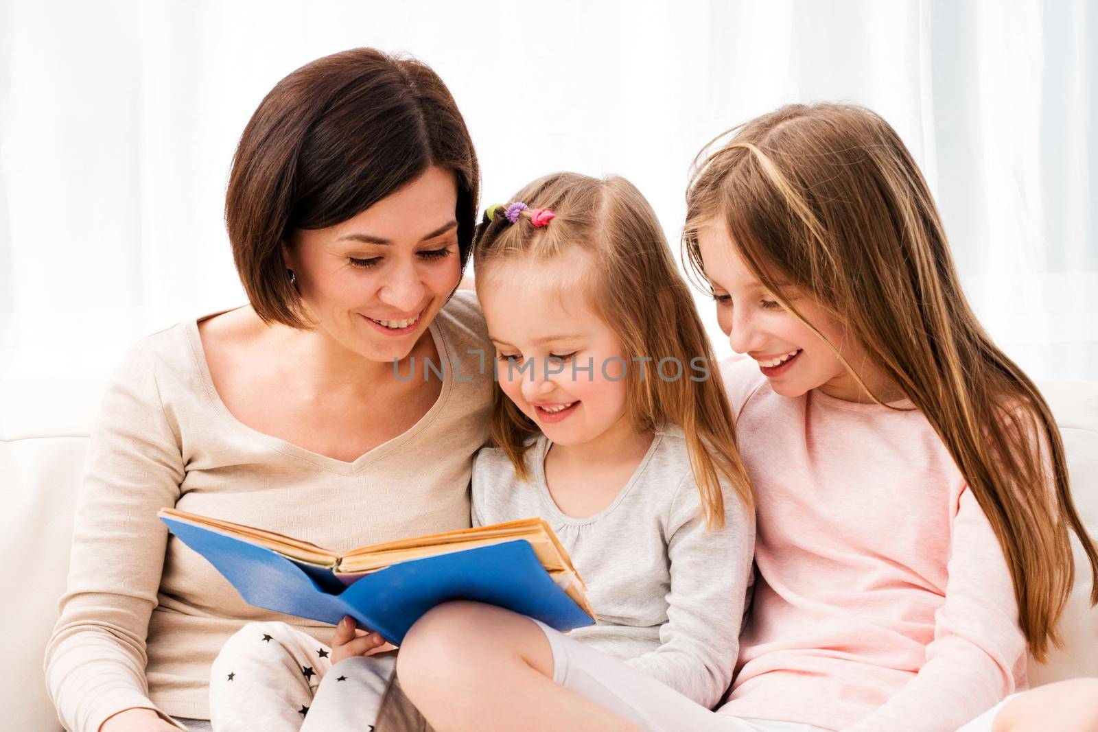 Mom and her two little daughters reading children's book in the living room. Mom and daughters sitting on the coach and reaading