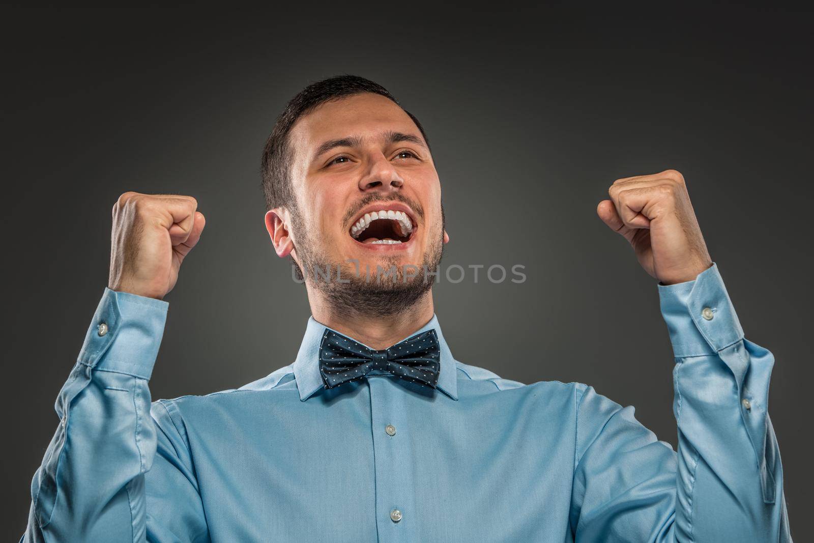 Closeup portrait of excited, energetic, happy, man in blue shirt and butterfly tie winning, arms up fists pumped celebrating success isolated grey studio background. Positive human emotion facial expression