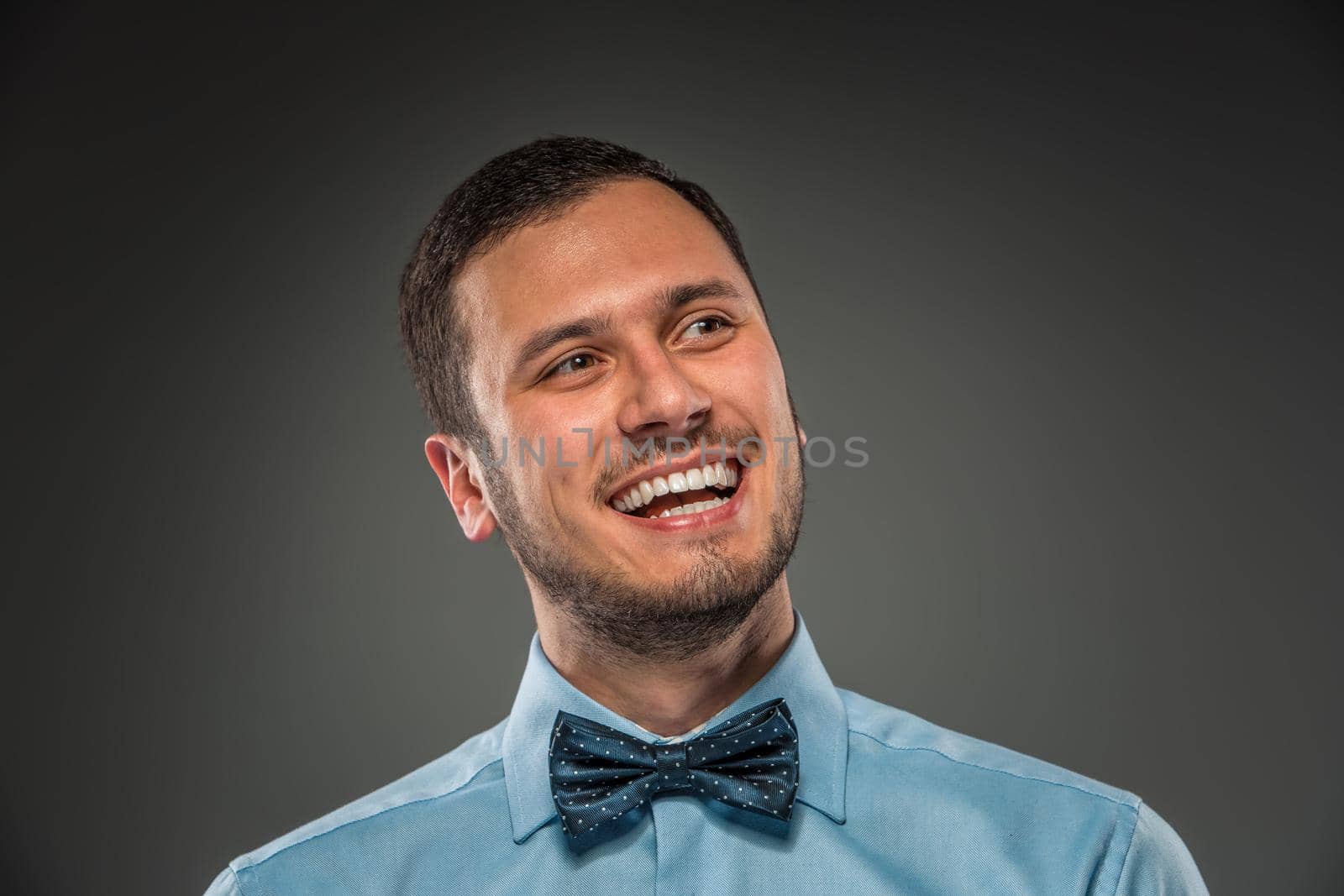 Portrait of happy smiling young man in blue shirt and butterfly tie isolated over gray studio background. Closeup