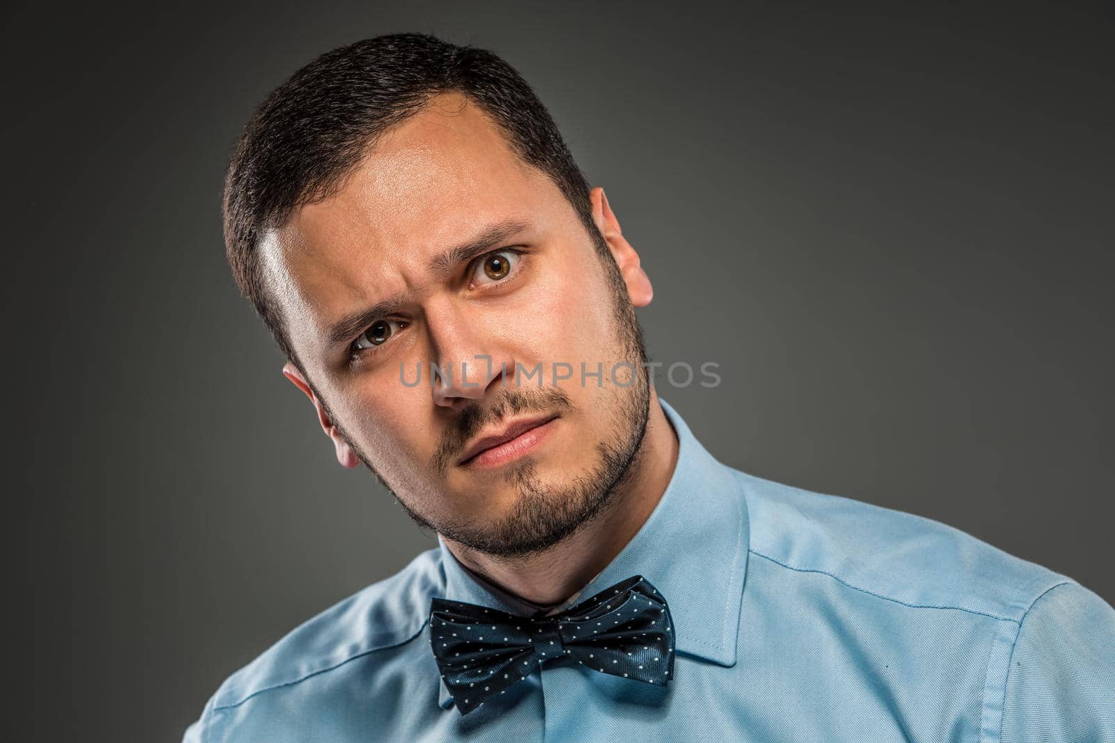 Portrait young man in blue shirt and butterfly tie looking at the camera with suspicion, isolated on gray studio background. Negative human emotion, facial expression. Closeup