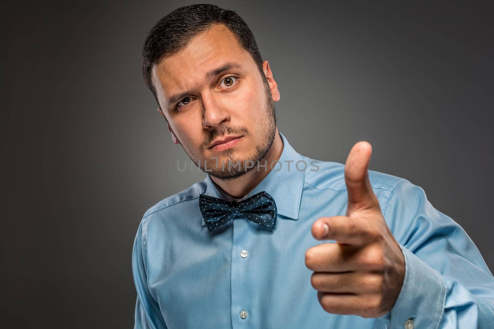 Closeup portrait of man in blue shirt and butterfly tie is gesturing with his hand, pointing finger at camera isolated on gray background.