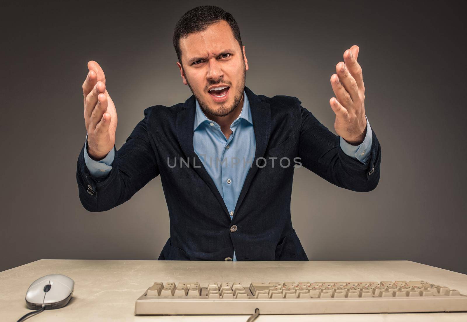 Portrait of angry upset young man in blue shirt and jacket with hands up yelling sitting at a desk near a computer isolated on gray background. Negative human emotion, facial expression. Closeup
