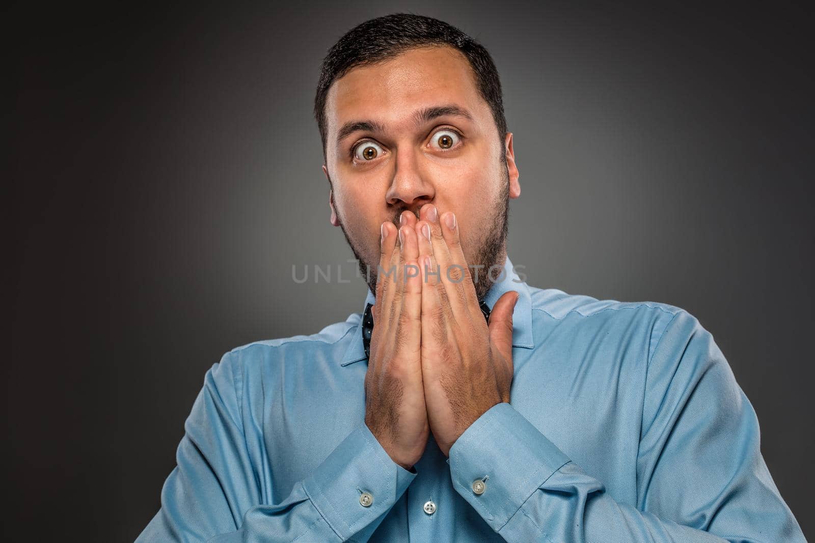 Portrait young man in blue shirt and butterfly tie looking at the camera with amazement, her hands covered her mouth isolated on gray studio background. Human emotion, facial expression. Closeup