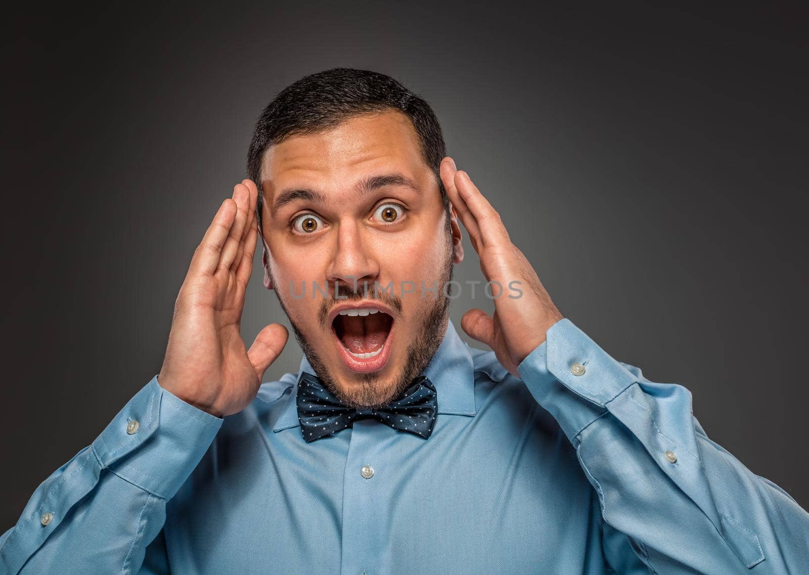 Portrait young man in blue shirt and butterfly tie looking at the camera with amazement, isolated on gray studio background. Human emotion, facial expression. Closeup