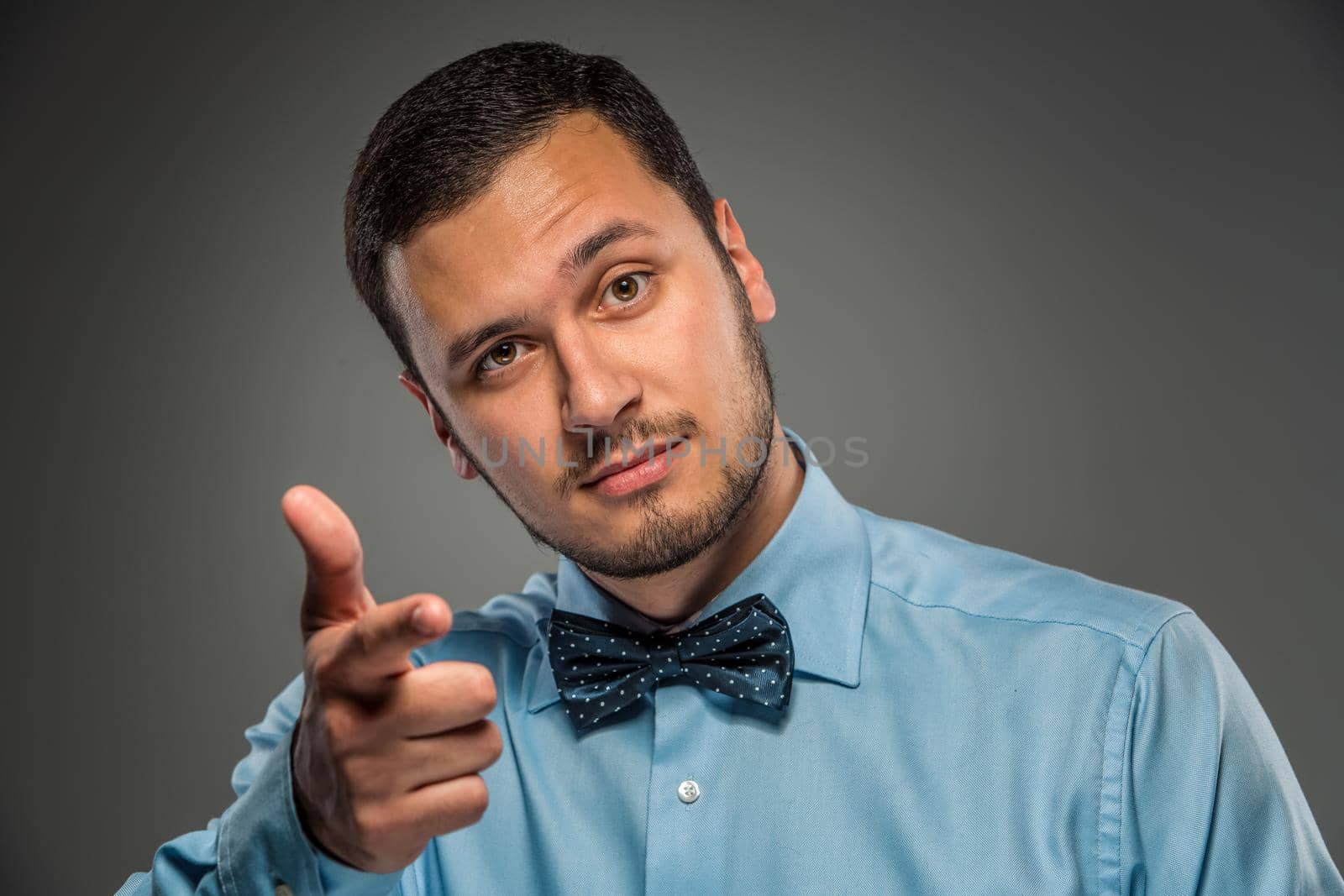 Closeup portrait of excited, energetic, happy, smiling man in blue shirt and butterfly tie is gesturing with his hand, pointing finger at camera isolated on gray background. Positive human emotion facial expression